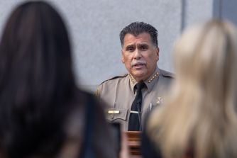 Robert Luna, Sheriff of the Los Angeles County Sheriff's Department, speaks at a podium during a briefing on the proposed DOJ settlement, with blurred figures in the foreground.