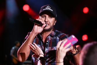 Luke Bryan performs on stage at a concert, wearing a plaid shirt and cap while singing into a microphone as fans reach toward him.