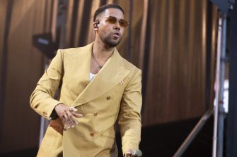 Romeo Santos wearing a yellow suit and sunglasses walks toward the camera.