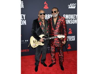 Two older men in matching black and red outfits with sunglasses pose on a red carpet, with one holding a guitar.