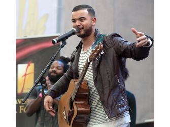 Guy Sebastian performing on stage with an acoustic guitar, singing into a microphone and gesturing with his right hand.