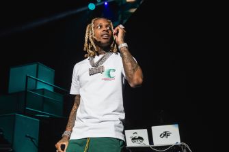 Lil Durk (Durk Banks) performs on stage in a white T-shirt and gold chains, microphone in hand.