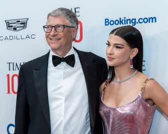 A gray-haired man in a black tuxedo and glasses stands beside a woman in a pink sequined dress on a red carpet at a formal event, with sponsor logos visible in the background.