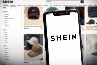 Smartphone screen shows the SHEIN logo in front of a blurred online marketplace page displaying hats and other clothing.