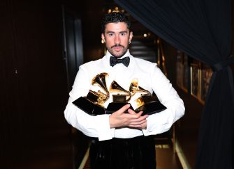 A man in a white tuxedo and black bow tie stands in a hallway, holding several gold trophy-like awards.