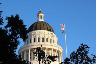 The California State Capitol dome in Sacramento, with flags flying on flagpoles in front and a clear blue sky.