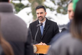 A man in a dark suit and tie speaks at a wooden podium with a microphone outdoors, the city seal visible on the podium, during a press event about New York City's pied-à-terre tax rollout.