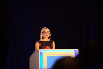 Kyrsten Sinema speaks at a podium on a stage, wearing glasses and a black and red sleeveless outfit.