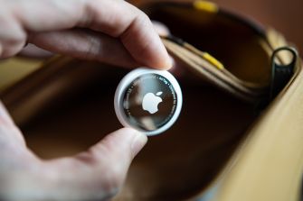 A hand holds a circular Apple AirTag with the Apple logo visible near the opening of a brown bag.
