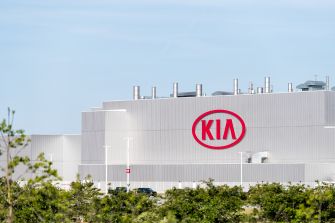 Kia Georgia manufacturing plant exterior with a prominent red Kia logo on a gray building and surrounding greenery under a blue sky.