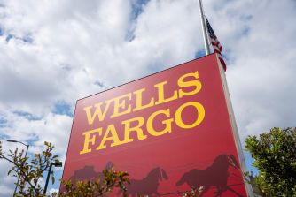Red Wells Fargo sign with yellow lettering and a mountain silhouette stands outdoors against a cloudy blue sky.