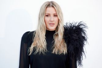 Ellie Goulding stands against a pale backdrop wearing a black top with a large black feathered shoulder detail.