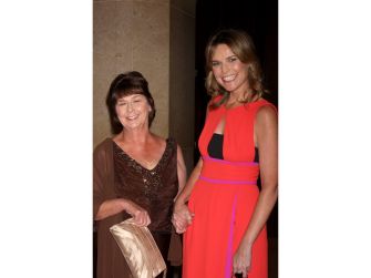 Two women stand side by side smiling for the camera, the older woman on the left wearing a brown top and holding a beige clutch, and the younger woman on the right wearing a bright orange dress with black accents.
