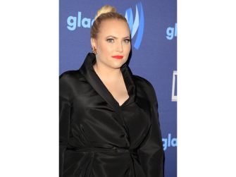 Meghan McCain, wearing a black satin blazer, poses for a photo at a formal event in front of a blue backdrop.