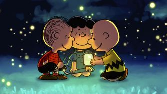 Three Peanuts characters sit in a circle on the grass at night, gathered around a glowing lantern under a starry sky.
