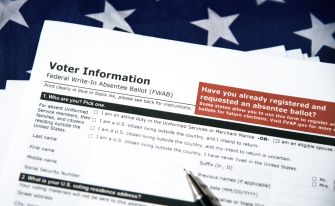 Close-up of voter information and absentee ballot forms, including a Federal Write-In Absentee Ballot (FWAB) heading, with a blurred American flag in the background.