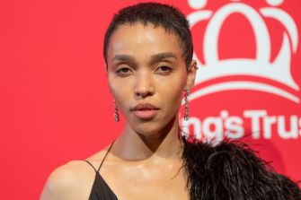 FKA Twigs, also known as Tahliah Barnett, poses in front of a red backdrop wearing dangling earrings.