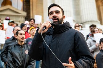 Zohran Mamdani, New York City mayor, speaks into a handheld microphone at a rally outside a government building with supporters in the background.