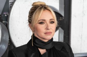 Hayden Panettiere with blonde hair styled in an updo, wearing a black outfit and scarf, gazes to the side.