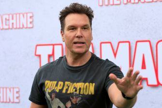 Dane Cook, wearing a black Pulp Fiction graphic T-shirt, gestures as he speaks at a promotional event.