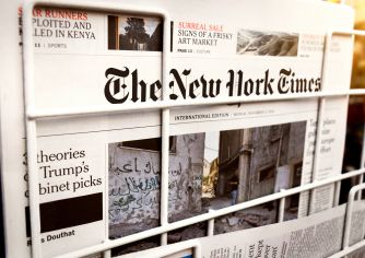 Front page of The New York Times displayed in a metal newspaper rack with other papers surrounding it.