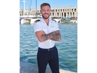 Nathan Gallagher stands with his arms crossed at a marina, wearing a white short-sleeve shirt and dark pants, with tattoos visible on his forearms.