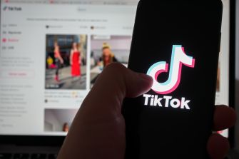 A hand holds a smartphone displaying the TikTok logo, with a laptop screen in the background showing a TikTok feed.