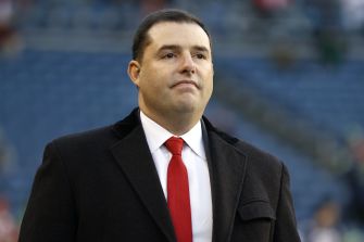 Jed York, principal owner of the San Francisco 49ers, wearing a dark overcoat and red tie, stands on a stadium sideline.