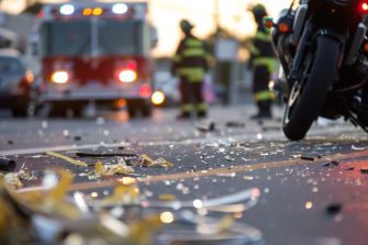 A motorcycle on a city street at an emergency scene, with an ambulance and firefighters in the background and debris scattered across the road.