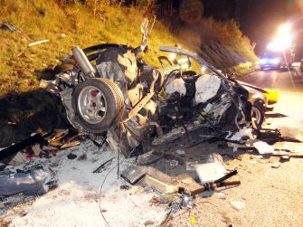 Nighttime road crash scene showing a severely damaged vehicle, with debris scattered across the pavement and emergency lights in the background.