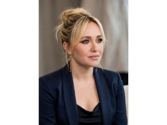 Hayden Panettiere seated in a navy blazer with a black top and her blonde hair in a loose updo, looking to the side.