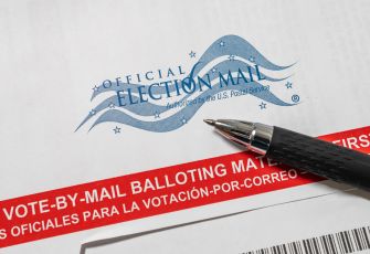 Close-up of an Official Election Mail stamp with a black pen resting on a ballot envelope, featuring a red banner reading "VOTE-BY-MAIL BALLOTING" to highlight mail-in voting.