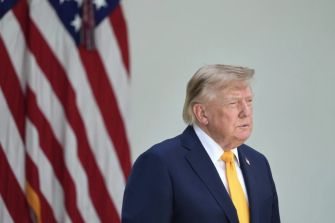 Donald Trump, wearing a navy suit and yellow tie, stands in front of American flags.