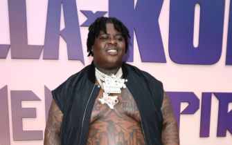 Rapper BigXthaPlug, real name Xavier Landum, poses at an event wearing an open vest and a heavy chain, with visible chest tattoos.