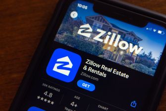 Close-up of a smartphone screen displaying the Zillow Real Estate & Rentals app page, featuring the Zillow logo, blue app icon, and a house image in the header.