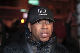 Russell Simmons wearing a black cap and dark puffer jacket, his mouth slightly open as he speaks.