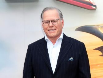 David Zaslav, CEO of Warner Bros. Discovery, stands in a dark pinstripe suit with a light blue shirt and pocket square, smiling in front of a backdrop.
