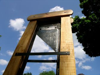Large wooden guillotine frame with a sharpened metal blade, photographed outdoors against a blue sky with trees in the background.