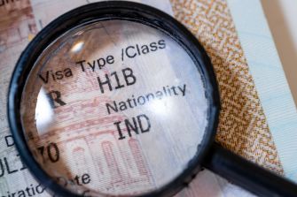 Magnifying glass over a visa document with "Visa Type H1B" and "Nationality IND" visible, highlighting immigration paperwork scrutiny.