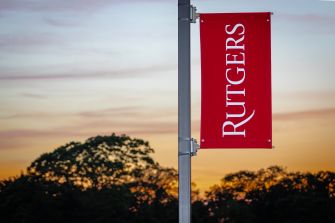 Rutgers University banner hanging on a flagpole with a sunset sky and trees in the background.