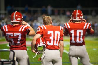 Three football players in red jerseys with white numbers stand with their backs to the camera on a football field, holding their helmets.