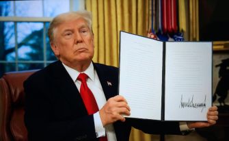 Donald J. Trump sits at a desk in the Oval Office, holding up an open, signed document.