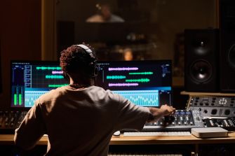 Person wearing headphones sits at a mixing console in a recording studio, adjusting the faders and controls of a digital audio workstation.