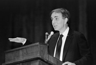 A man in a dark suit and tie stands at a wooden podium, gesturing with his left hand while speaking into a microphone.