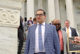 George Santos, wearing a blue checkered suit and glasses, walks down steps from a building with onlookers nearby.