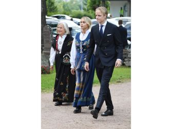 Three people walk outdoors near a royal estate: an older woman in traditional dress, a younger woman in a blue-patterned outfit, and a man in a dark suit.