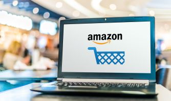 Laptop on a table displays the Amazon logo with a blue shopping cart icon against a blurred store background.