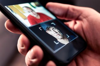 Close-up of a hand holding a smartphone displaying a grid of AI-generated intimate images, with a rectangular focus frame framing a person's face in the foreground.