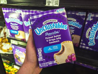 A hand holding a purple box of Smucker’s Uncrustables peanut butter and grape jelly sandwich, displayed on a grocery-store shelf.