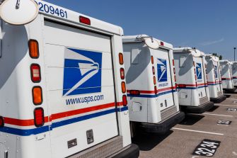 Row of United States Postal Service delivery trucks with logos parked in a lot.